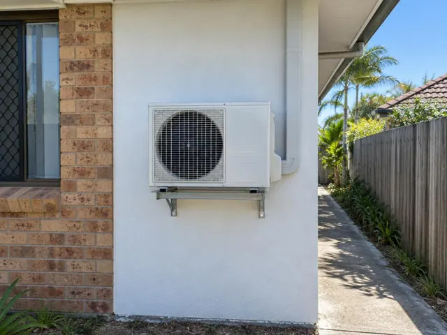 Wall-mounted outdoor air conditioner condenser on a rendered exterior wall beside a brick house in Burleigh Heads